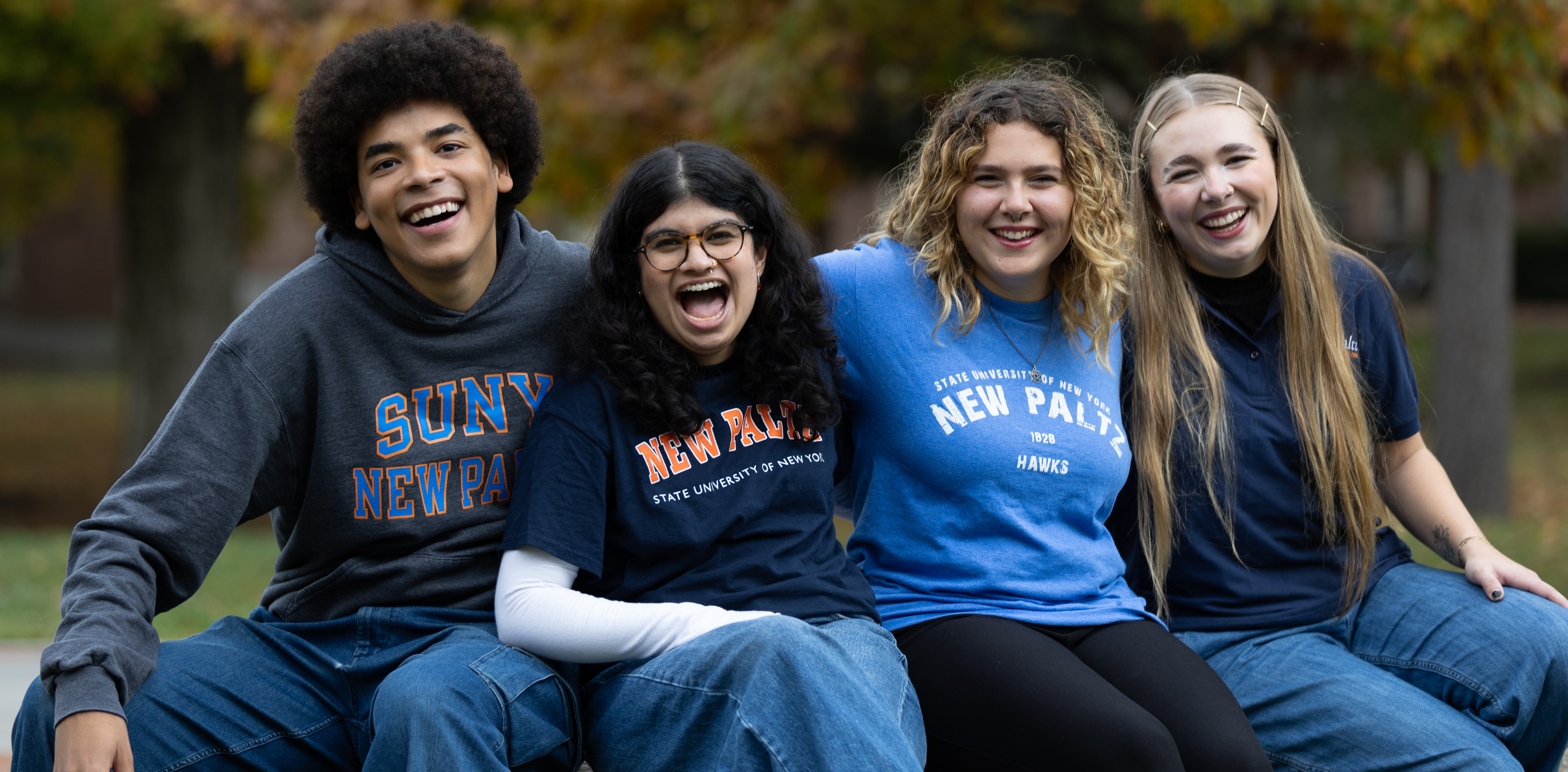 Four Students sitting side by side smiling wearing New Paltz Shirts